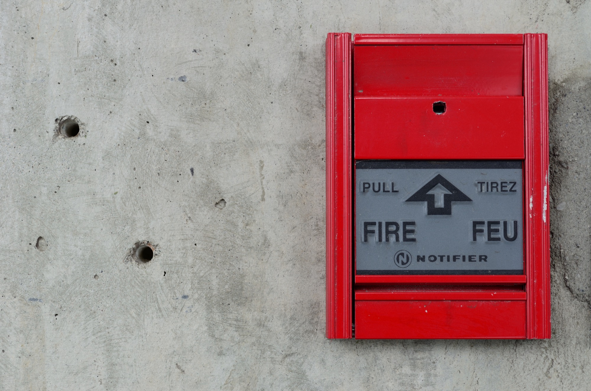 A red manual fire alarm call point mounted on a bare concrete wall.